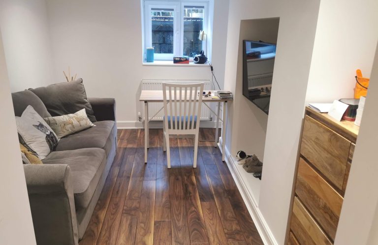 Small, modern room with a grey sofa, desk, and wooden dresser. Natural light from a window.