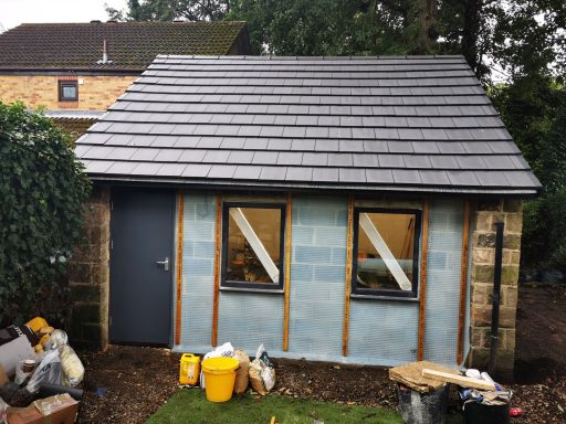 A small building with a sloped roof, two windows, and construction materials outside.