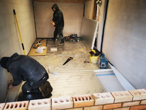 Two workers are constructing a room, laying flooring and working on walls.