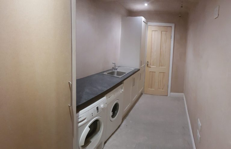 Narrow laundry room with appliances and a wooden door at the far end.