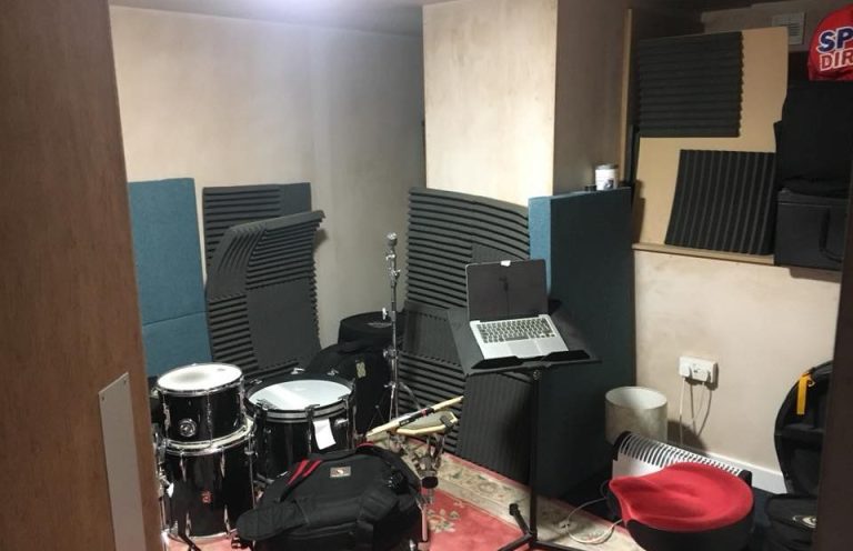 Music rehearsal space with drums, music stand, laptop, and soundproofing panels.