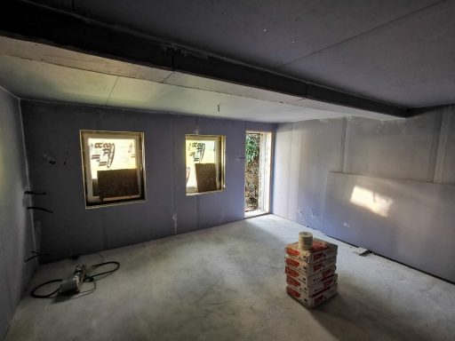 Empty room with three windows, a doorway, and a stack of bricks in the centre.