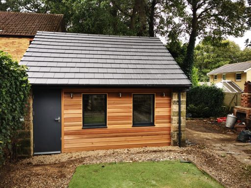 Small wooden garden building with a sloped roof and large windows. Surrounded by trees.